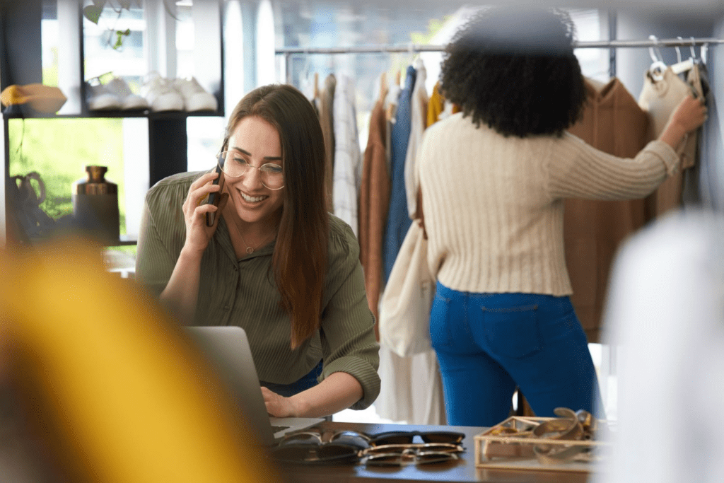 A woman in a clothing boutique smiles while talking on a smartphone and using a laptop, with another person browsing clothes in the background. This image highlights how retail businesses use VoIP phone services and cloud-based phones for cost-effective communication and customer support.