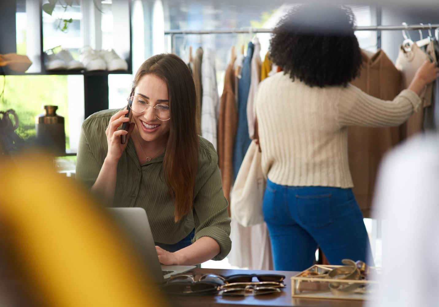 A woman in a clothing boutique smiles while talking on a smartphone and using a laptop, with another person browsing clothes in the background. This image highlights how retail businesses use VoIP phone services and cloud-based phones for cost-effective communication and customer support.