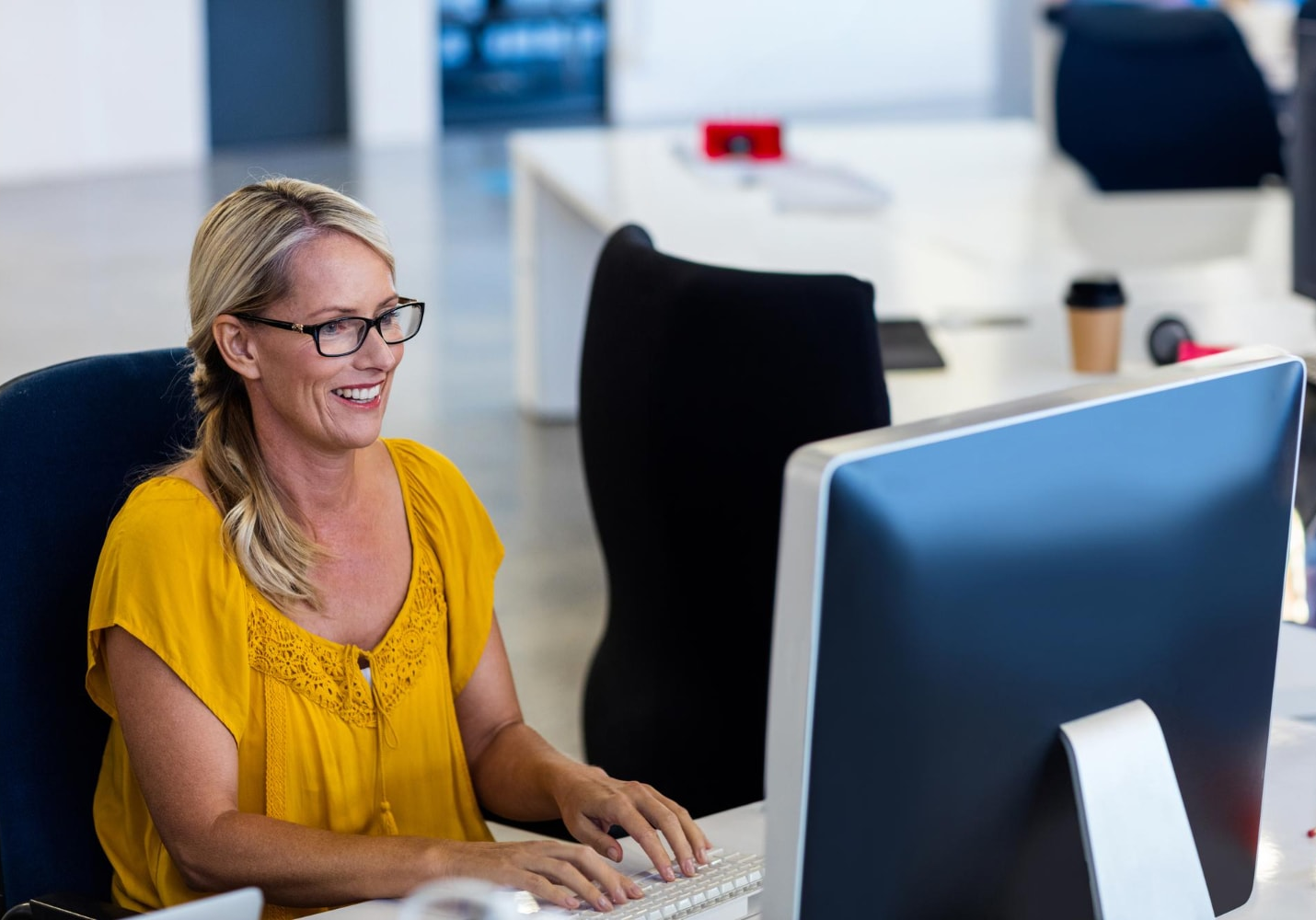 Smiling woman in a yellow blouse working on a desktop computer in a bright, modern office, exemplifying efficient business communication with upgraded business phone systems in a tech-driven environment.