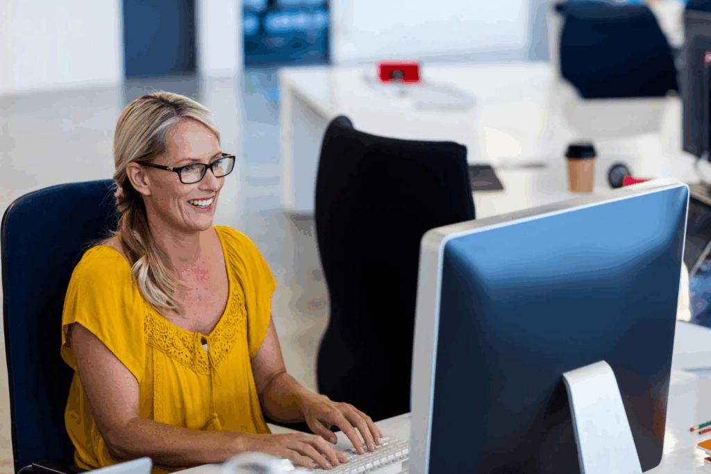 Smiling woman in a yellow blouse working on a desktop computer in a bright, modern office, exemplifying efficient business communication with upgraded business phone systems in a tech-driven environment.